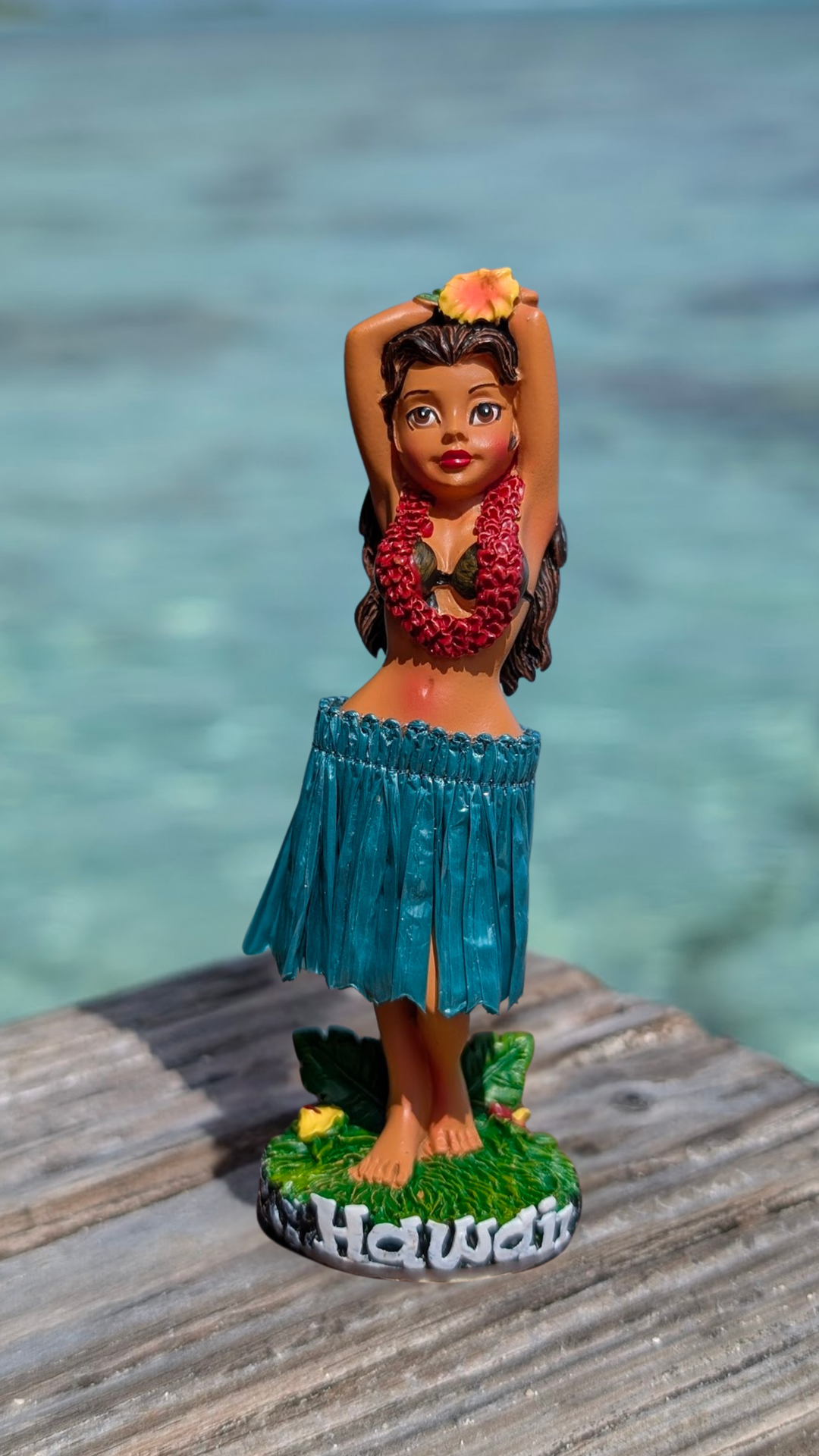 A figurine of a hula girl wearing a hula skirt and holding a flower, standing on a wooden dock with the word "Hawaii" written on it.