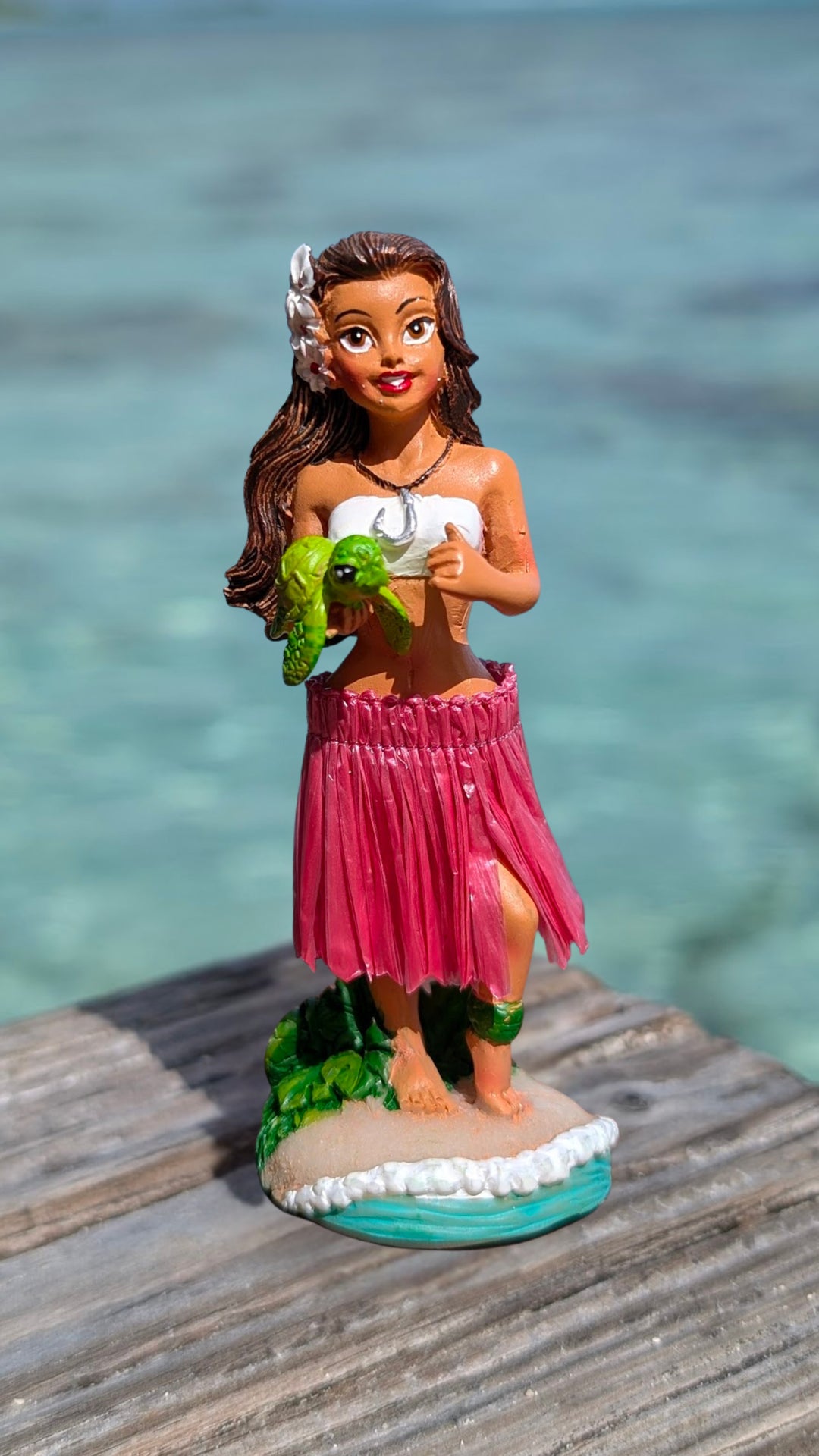 A dashboard doll figurine of a hula girl wearing a pink skirt and holding a green turtle