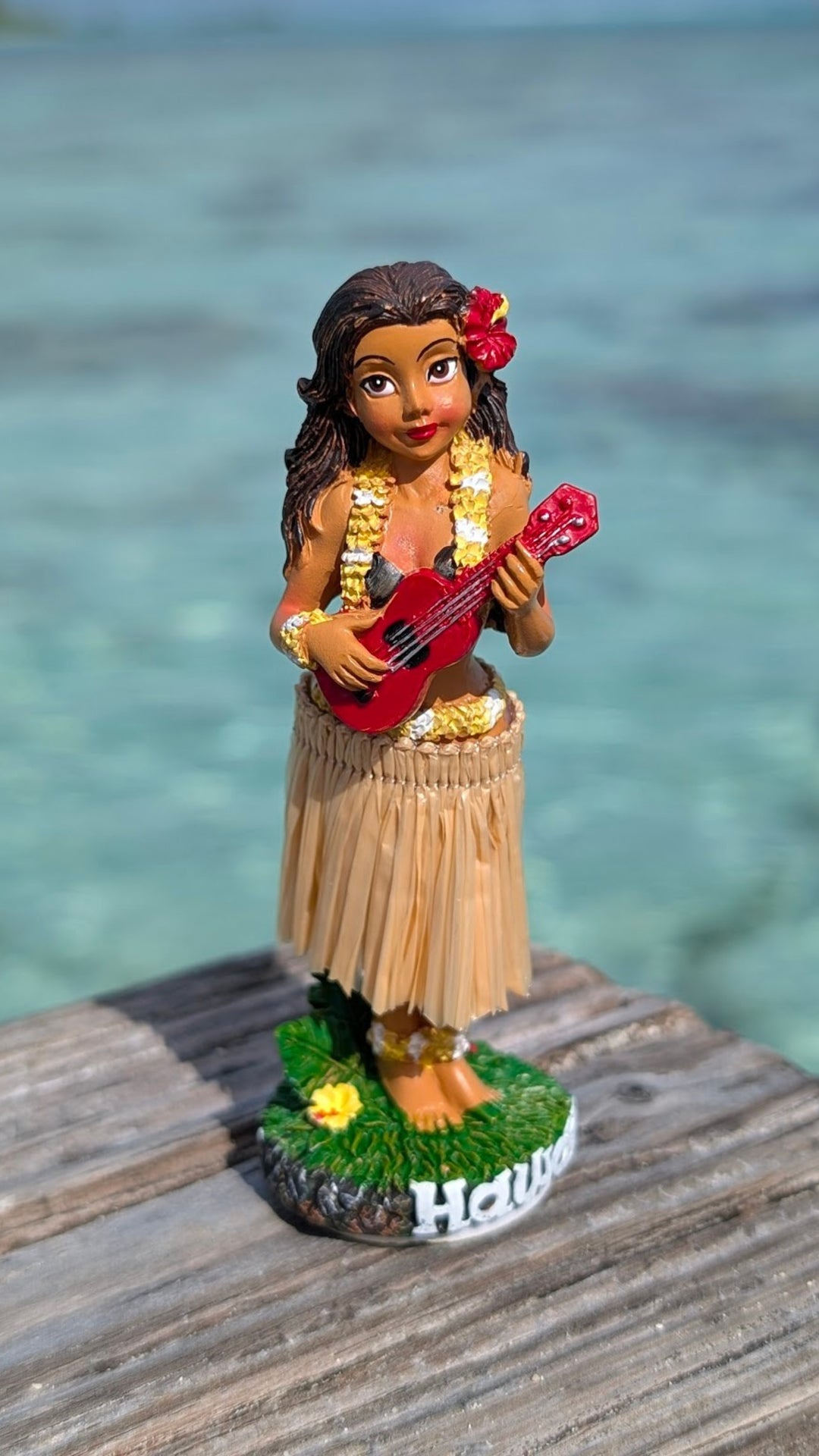 A dashboard doll figurine of a woman playing a red guitar, wearing a hula skirt, and standing on a wooden platform with the word "Hawaii" written on it.