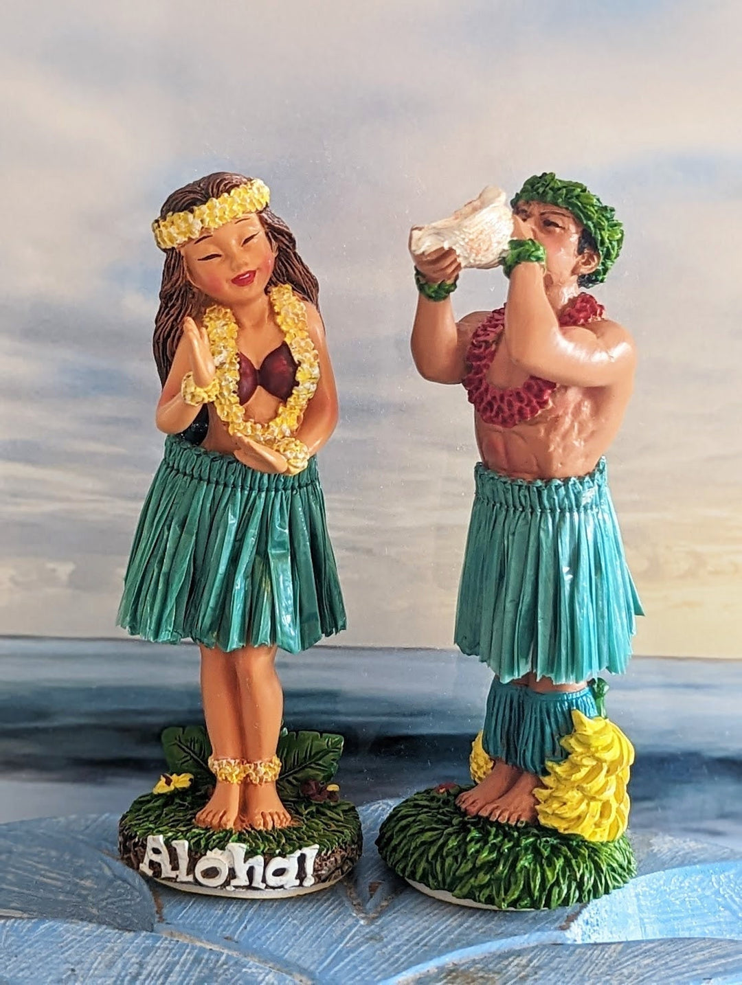 Two figurines of a man and a woman dressed in traditional Hawaiian attire, with the word "Aloha" written on them, standing on a grassy surface against a cloudy sky background.