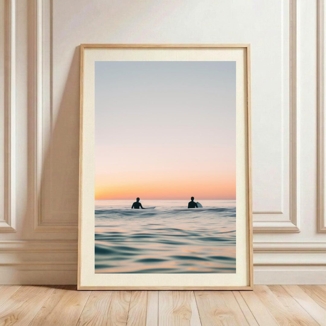 A minimalist watercolor fine art print showing a tranquil ocean scene at sunset. Two surfers are silhouetted sitting on their surfboards in the water. The sky is a gradient of soft blue, pink, and warm orange, which is beautifully reflected on the glassy surface of the ocean.