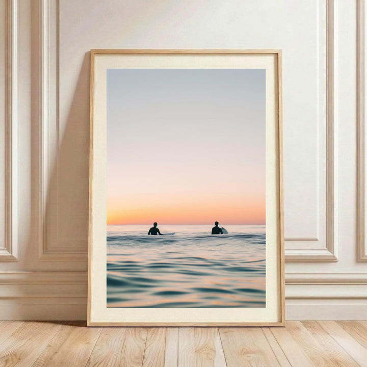 A minimalist watercolor fine art print showing a tranquil ocean scene at sunset. Two surfers are silhouetted sitting on their surfboards in the water. The sky is a gradient of soft blue, pink, and warm orange, which is beautifully reflected on the glassy surface of the ocean.